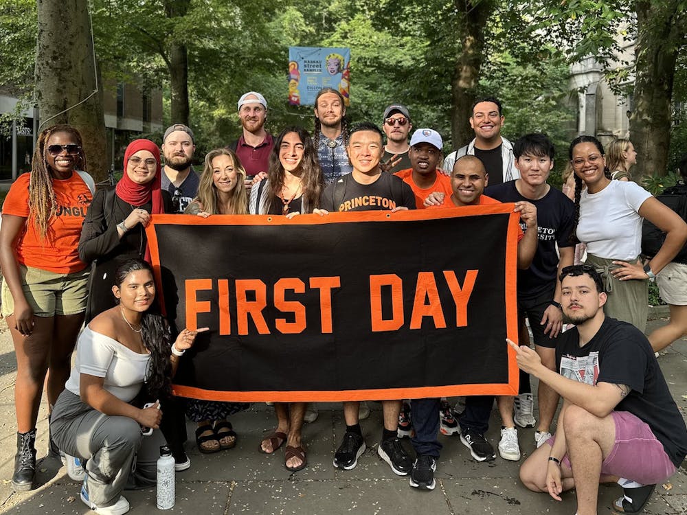 A group of transfer students stands by McCosh hall holding a flag that says "First Day" and smiling.