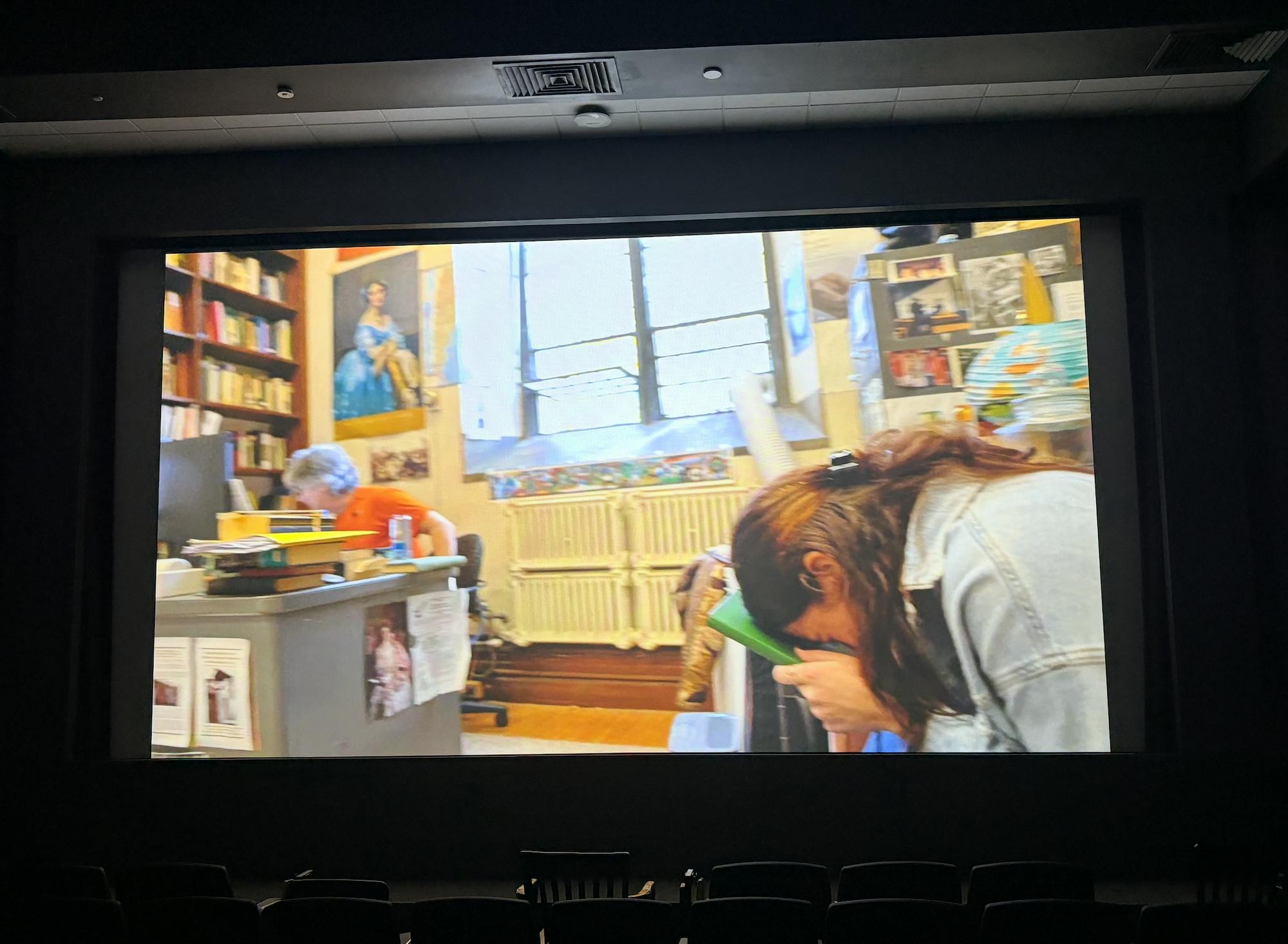 A colorful office contains two people. A light-skinned man wearing an orange shirt is sitting at a desk. A girl with brown hair and light-skin is wearing a blue jean jacket. She is in a crunched over position holding a green book to her forehead.