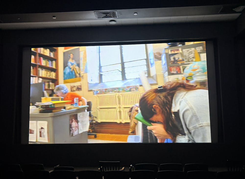 A colorful office contains two people. A light-skinned man wearing an orange shirt is sitting at a desk. A girl with brown hair and light-skin is wearing a blue jean jacket. She is in a crunched over position holding a green book to her forehead.
