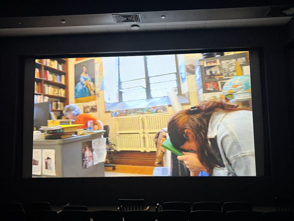 A colorful office contains two people. A light-skinned man wearing an orange shirt is sitting at a desk. A girl with brown hair and light-skin is wearing a blue jean jacket. She is in a crunched over position holding a green book to her forehead.