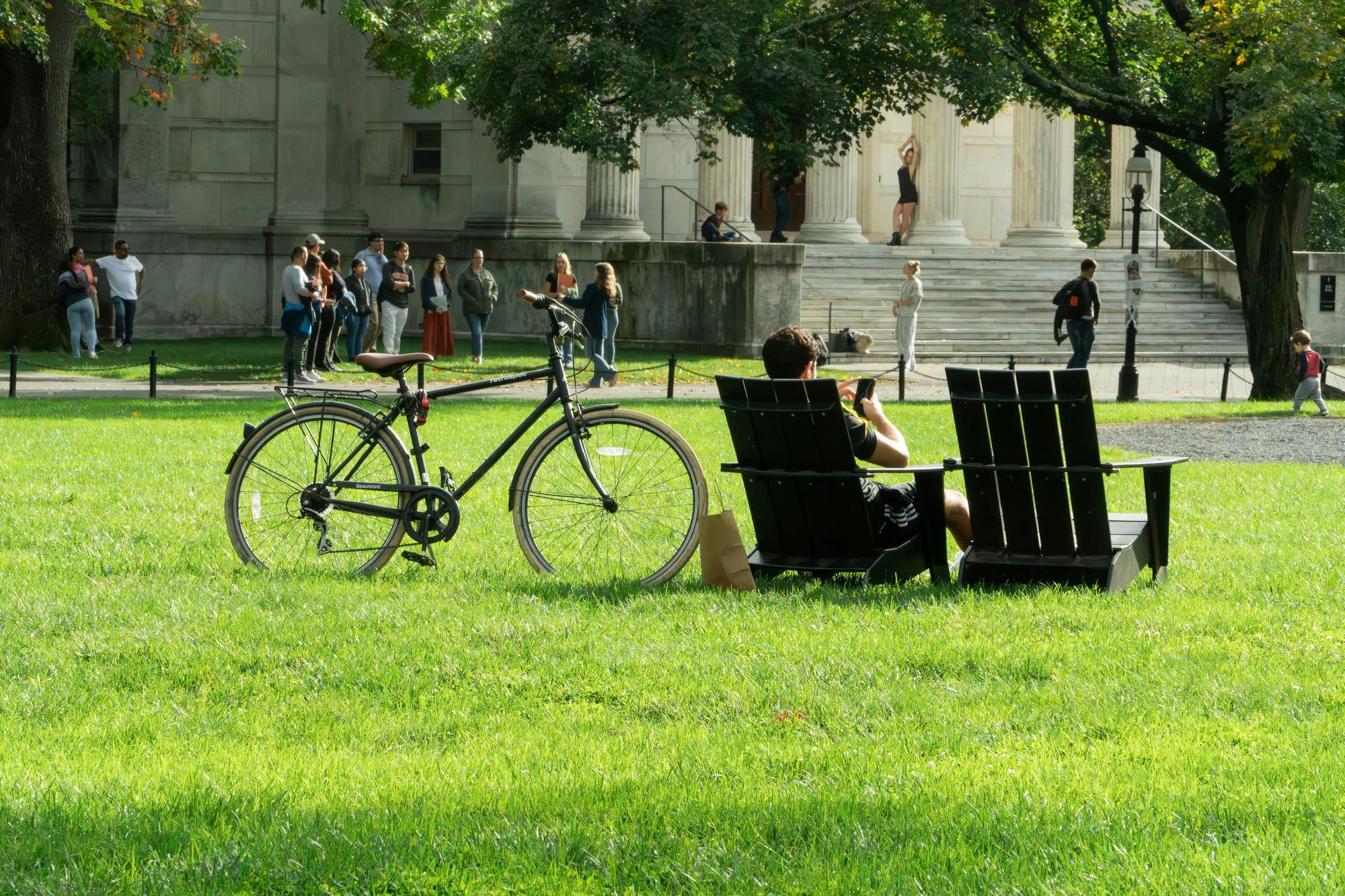 A student is sitting on a chair in Cannon Green with his bike next to him. 