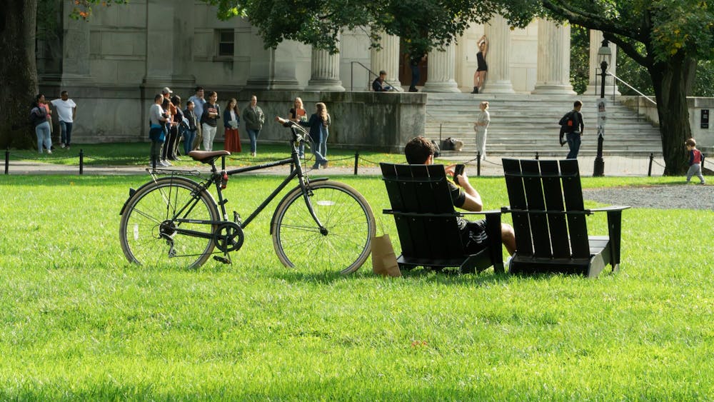 A student is sitting on a chair in Cannon Green with his bike next to him. 