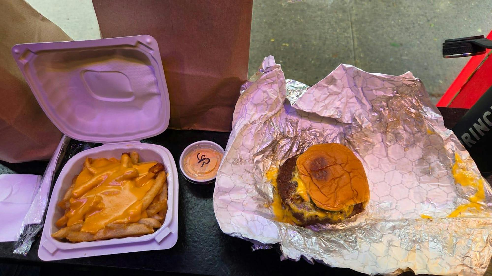 There is a gray piece of foil with a brown bun and a black burger patty inside. There is yellow cheese splattered on the patty. There is a small white to-go container that has brown fries with yellow cheese on top.