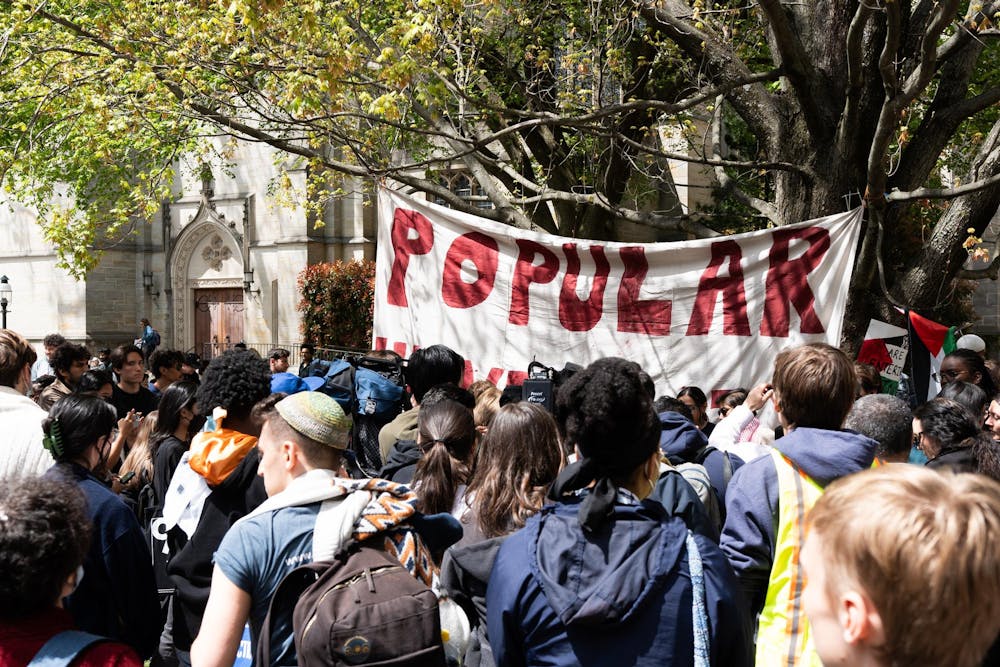 A group of students standing in McCosh courtyard facing a side door of the University Chapel, in front of a white sign hanging from a tree where the word "Popular" is written in all-capital red letters.