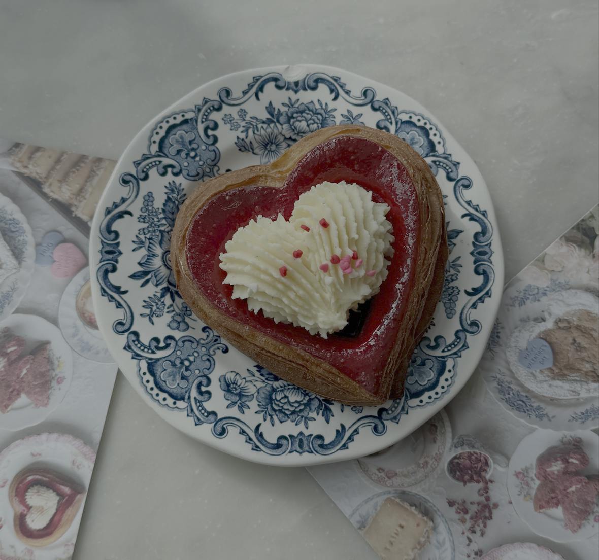 A heart-shaped flaky croissant pastry with cream piped and heart-shaped pink and red sprinkles on top sits on a plate with blue floral designs. 