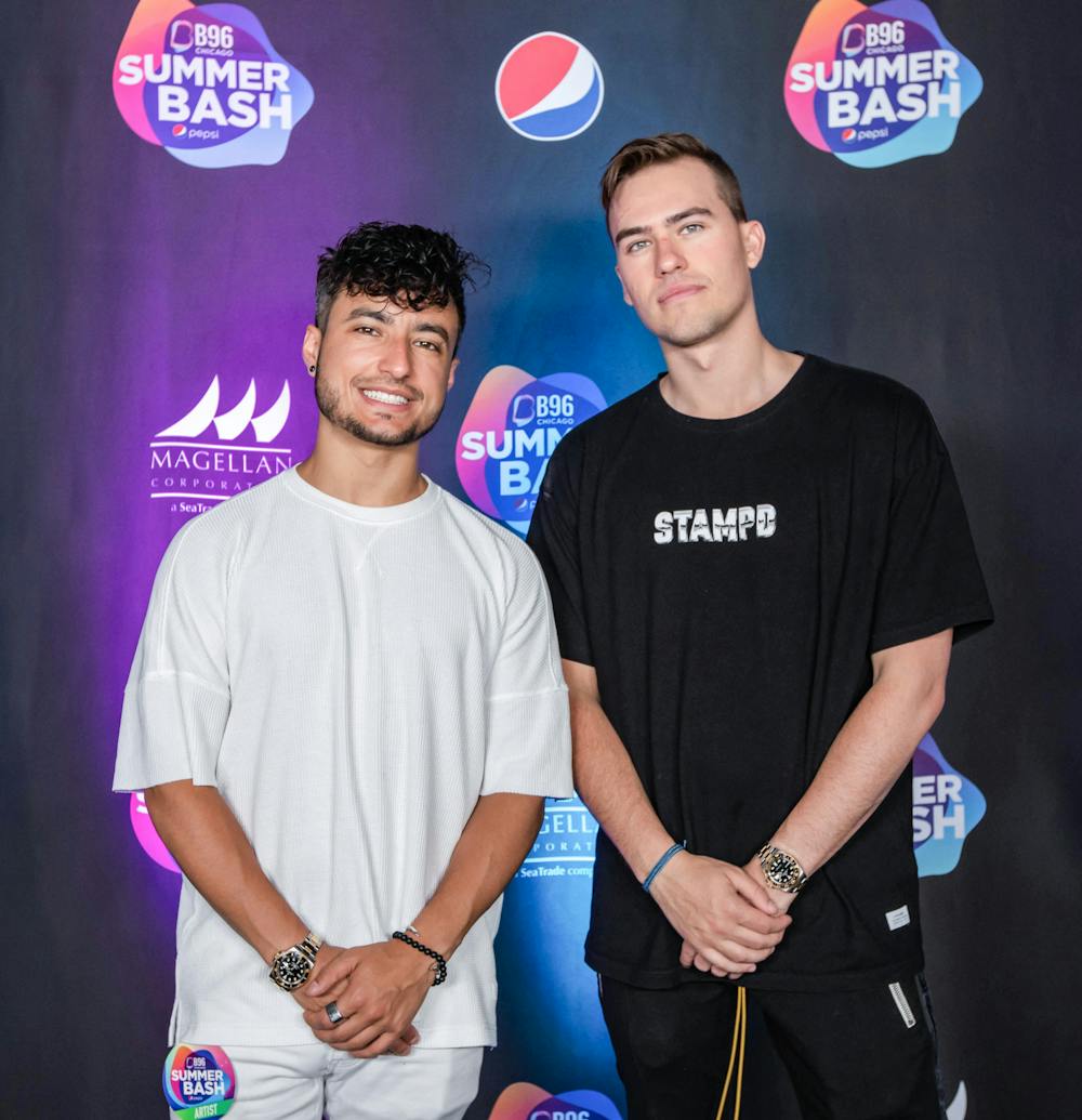 Two men, one shorter with dark hair and one taller with light hair, stand in front of a background for the B96 Chicago Summer Bash