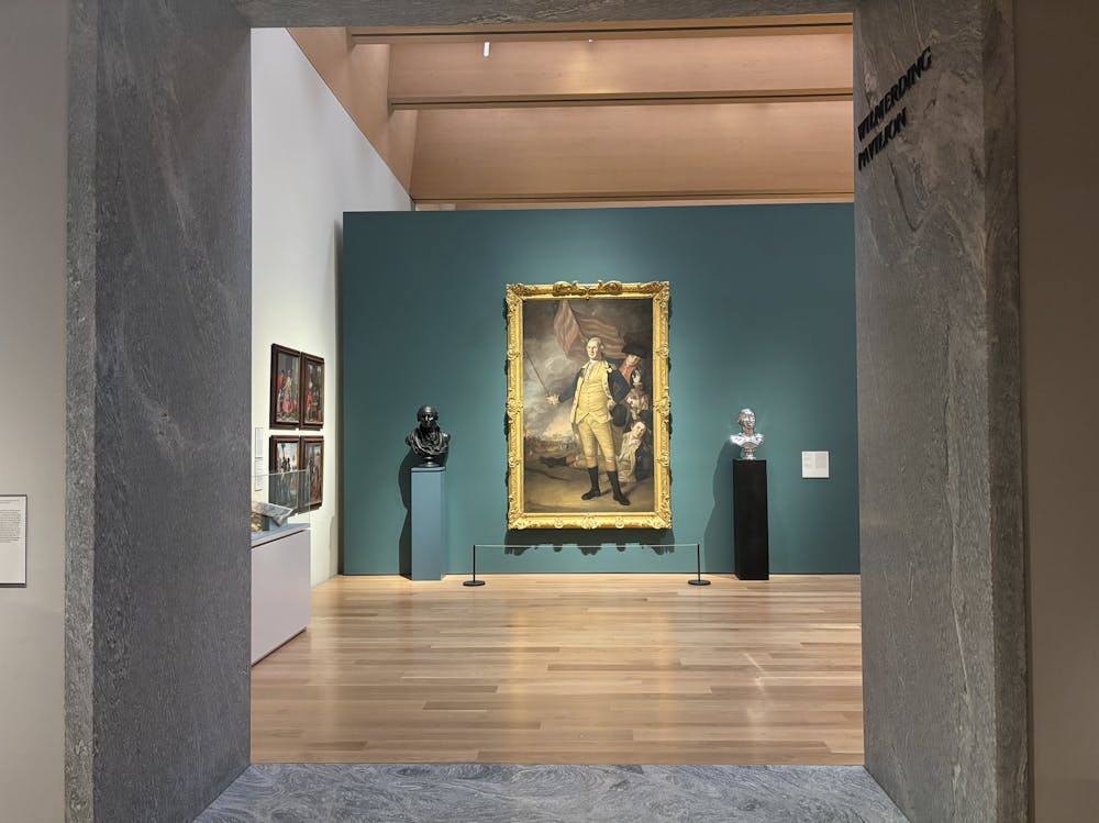 A work depicting George Washington in a white and blue outfit is hanging on a teal blue wall. It is sandwiched by two busts, one is black and one is silver.