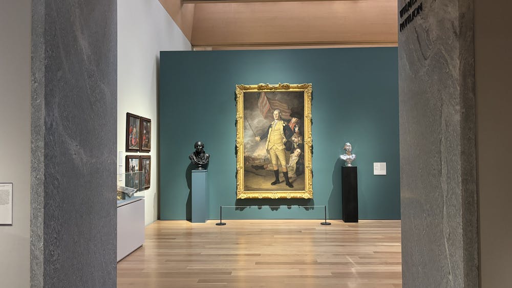 A work depicting George Washington in a white and blue outfit is hanging on a teal blue wall. It is sandwiched by two busts, one is black and one is silver.