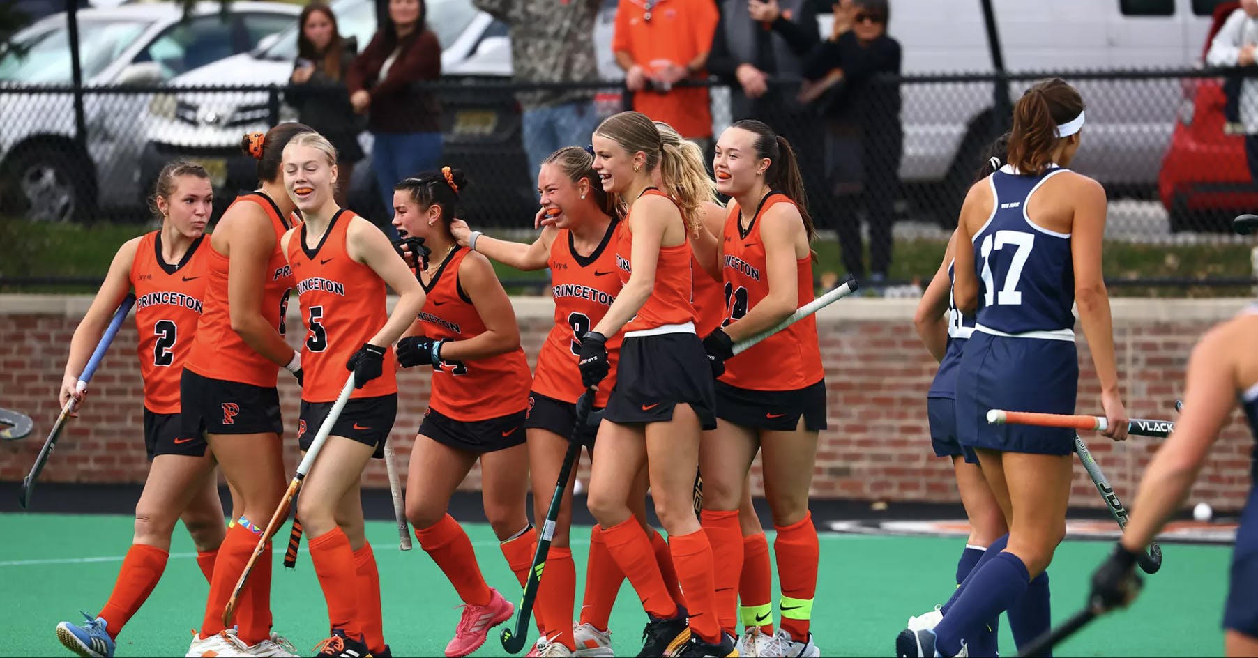 Eight field hockey players in orange tank tops, black shorts, and orange socks smile as they walk off the field.