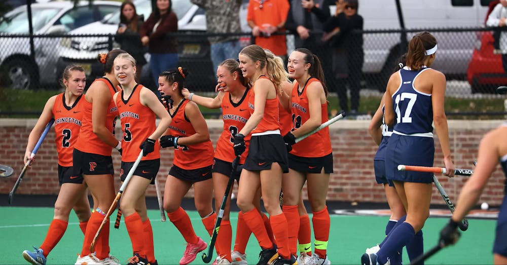 Eight field hockey players in orange tank tops, black shorts, and orange socks smile as they walk off the field.