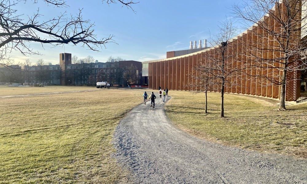 A la derecha, un edificio rojizo con faldones metálicos para dar sombra, tras un camino de grava con varios ciclistas. Césped a la izquierda.