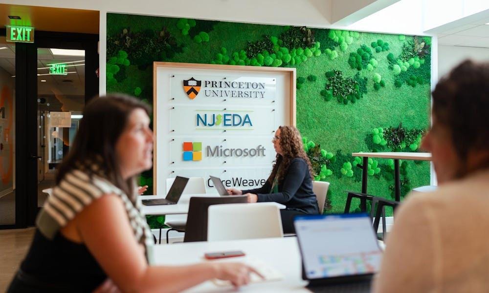 Enfocó en una mujer trabajando en su computadora, sentada enfrente de un signo del “AI Hub Founding Partners”. 