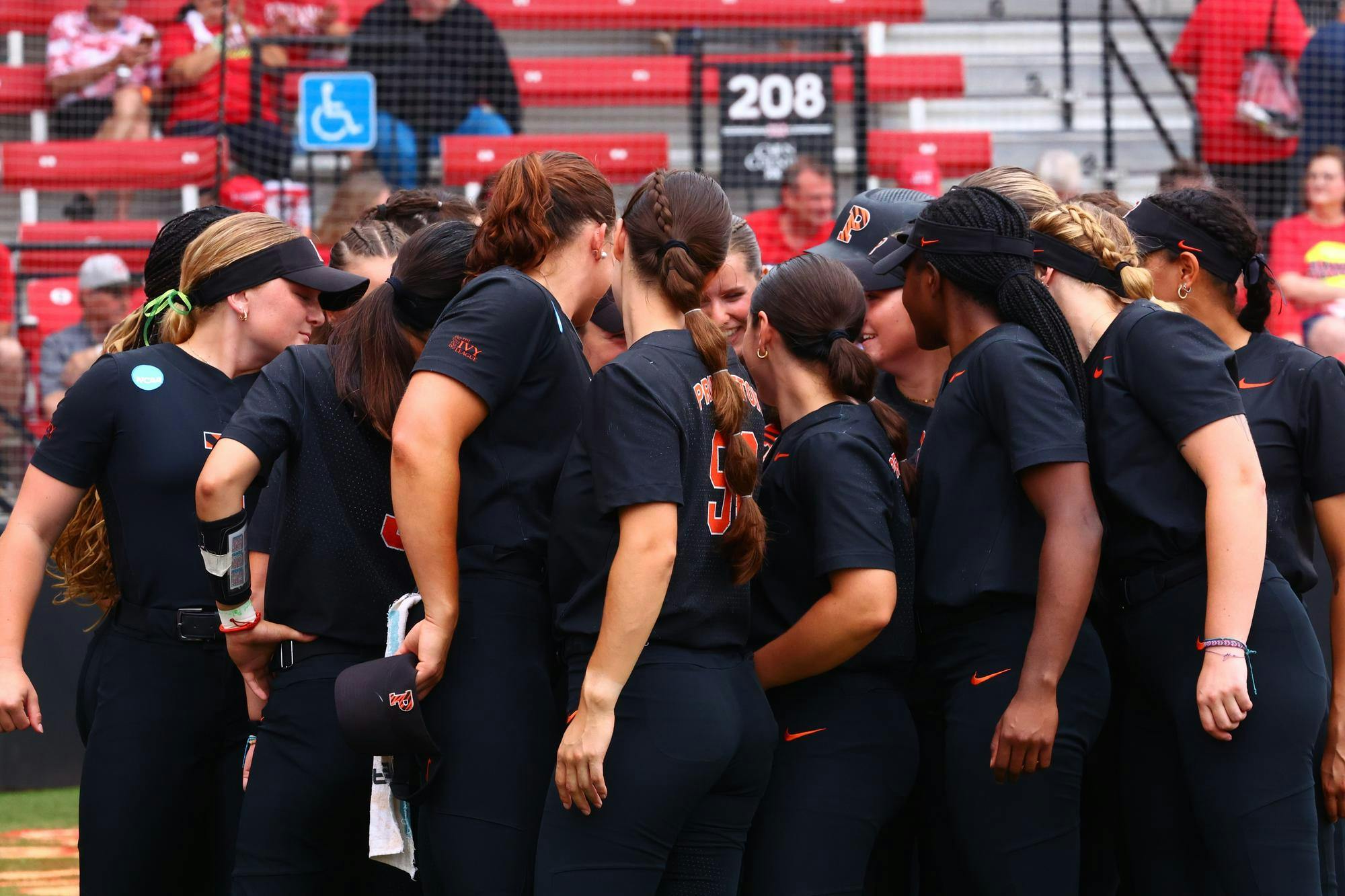 Group of women in black shirts with orange lettering in a circle in front of red bleachers.