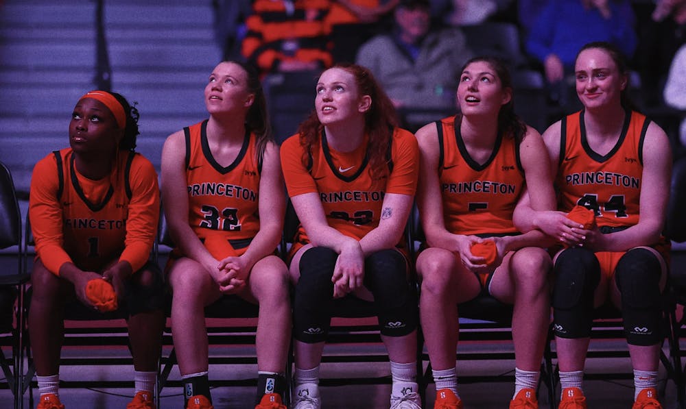 Five women's basketball players in orange jerseys sit on the bench, looking up.