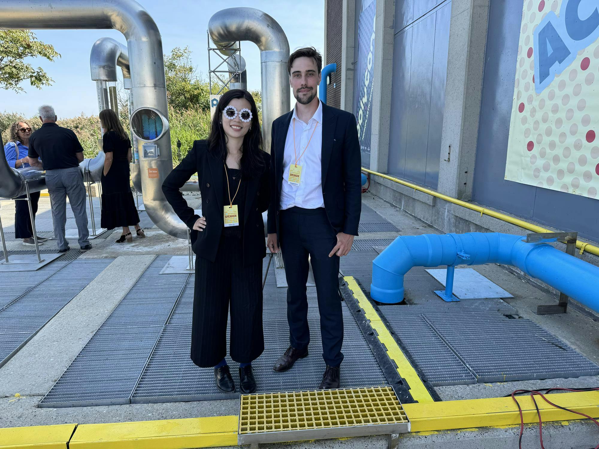 Yidian Liu and Nathaniel Banks stand side-by-side at an outdoor site with large pipes in the background.