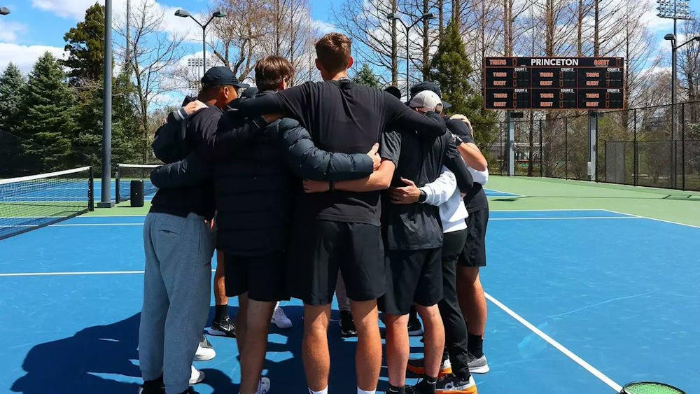 A group of men huddle together on a tennis field surrounded by trees.