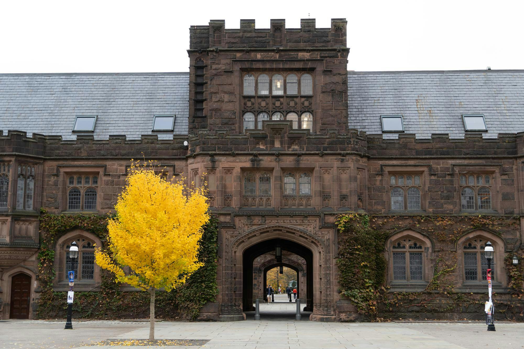 Foreground: yellow and green leafed tree with lamp posts on either side. Background: brown building with large arch in center. 2-story tower above arch. 