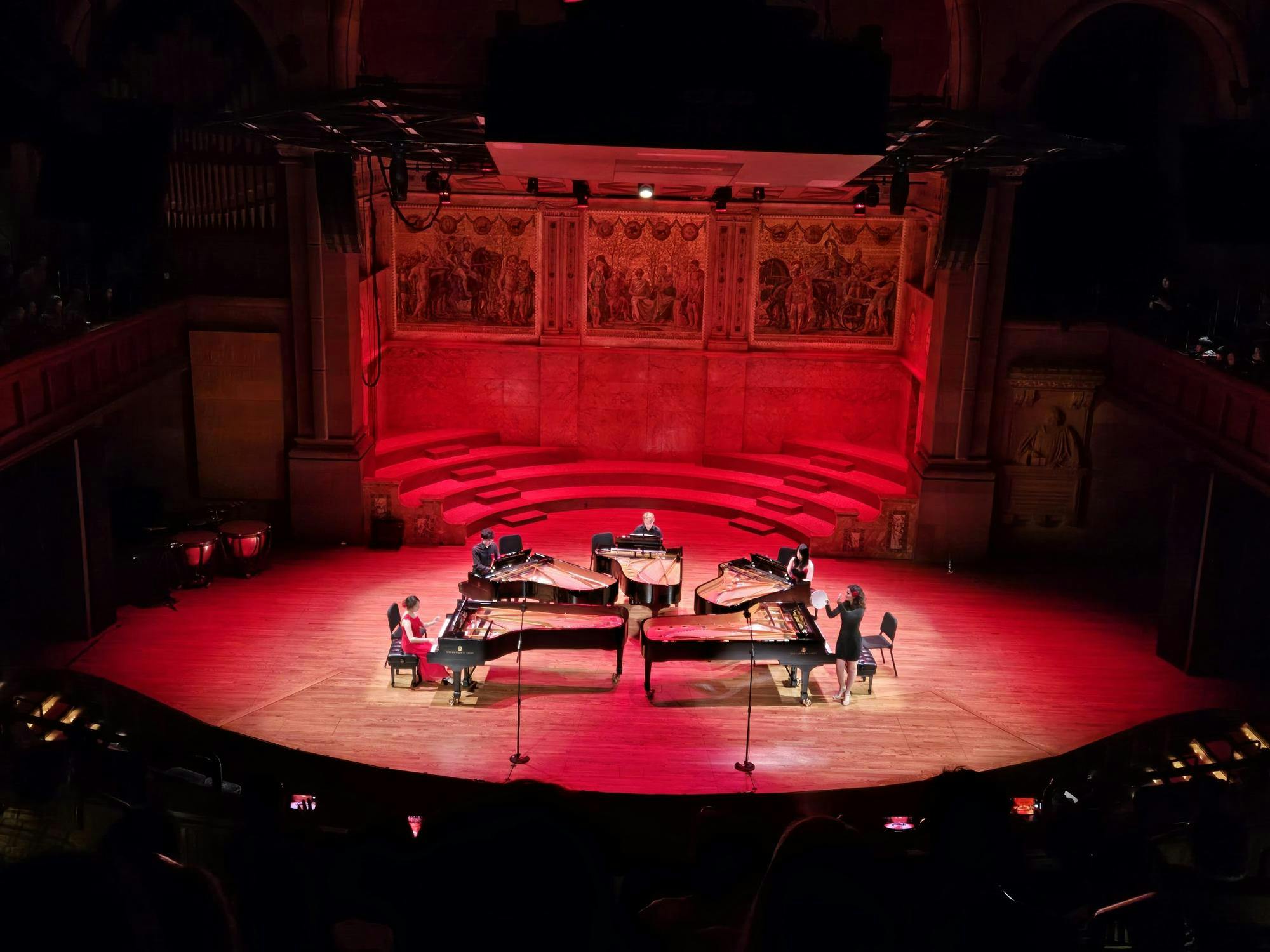 Red light illuminates a stage with five pianos.
