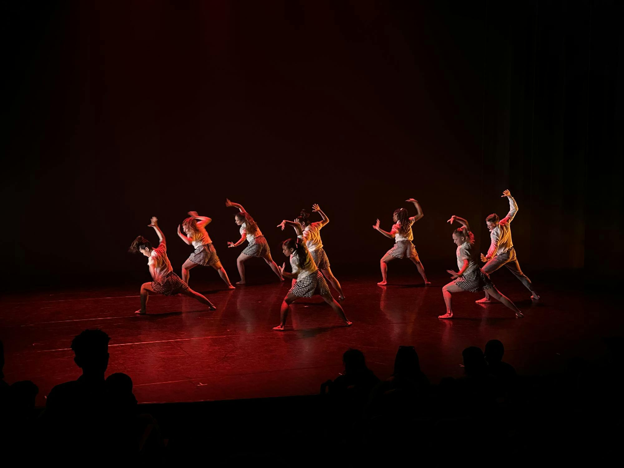 Dancers pose, lunging to the left with their hands in the air, on a black stage illuminated by red lighting.