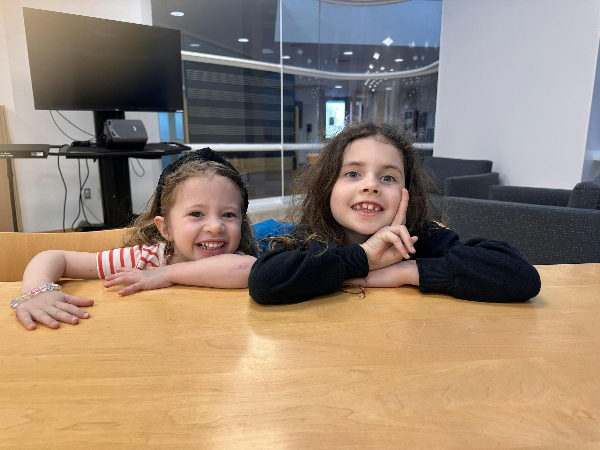 Two girls pose and smile at the camera from behind a table with couches and a computer in the background.