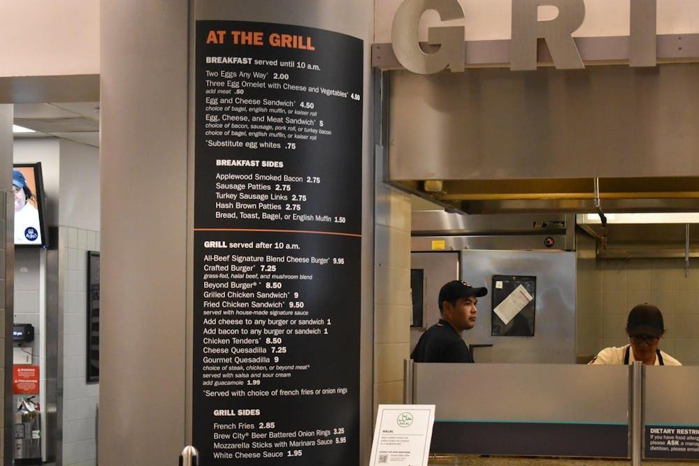 A menu with a black background and white writing pasted on a pillar. There are people in black and white clothes in the background.