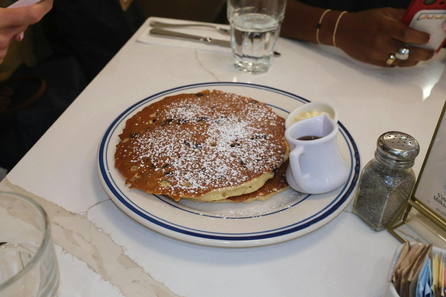 There is a white plate with a blue rim on top of a white marble-patterned table. The table has black pepper and salt shaker. There are brown pancakes with small dark brown chocolate chips in them. The pancakes are covered in white powdered sugar. There are also two small white pitches with brown sauce and light yellow butter.