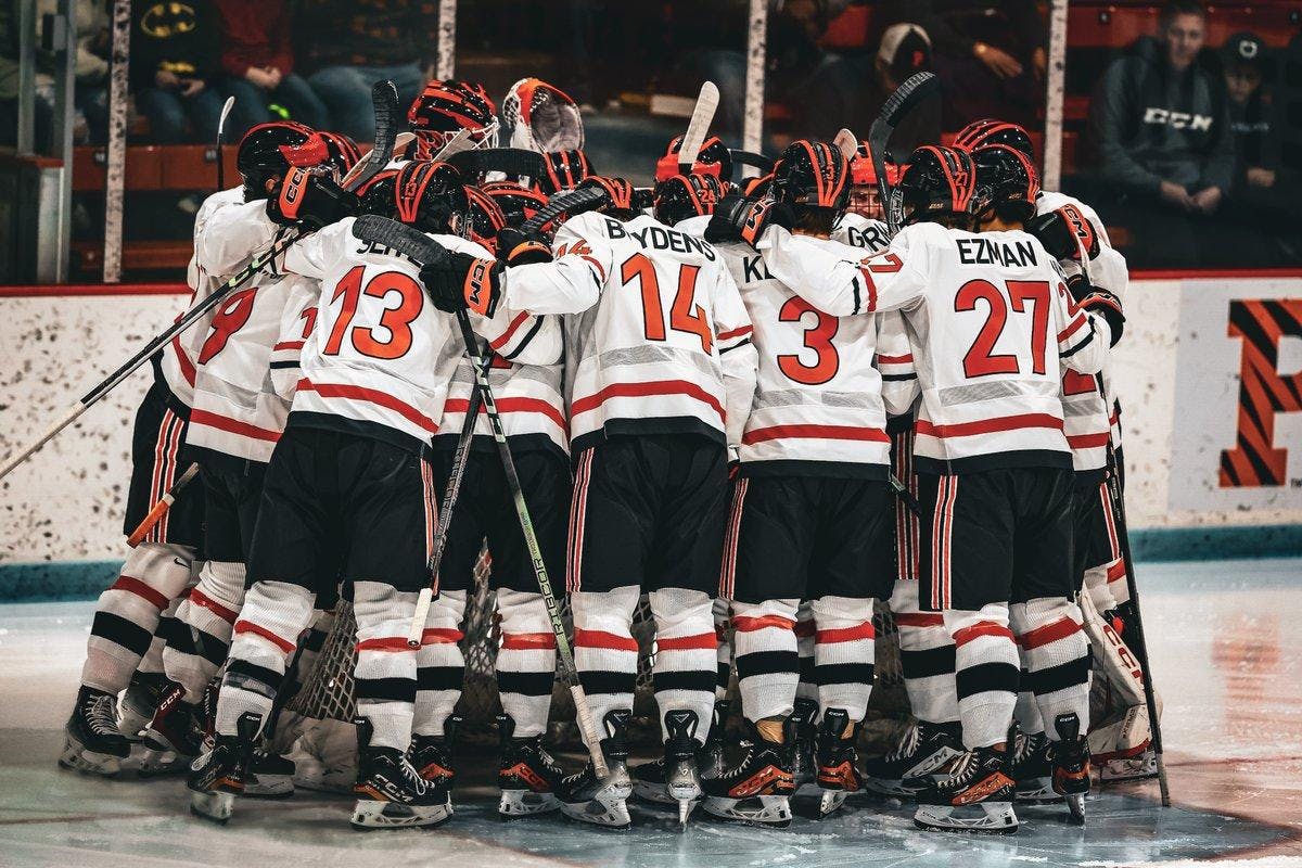 Men in orange and white hockey uniforms gather in group on ice surface. 