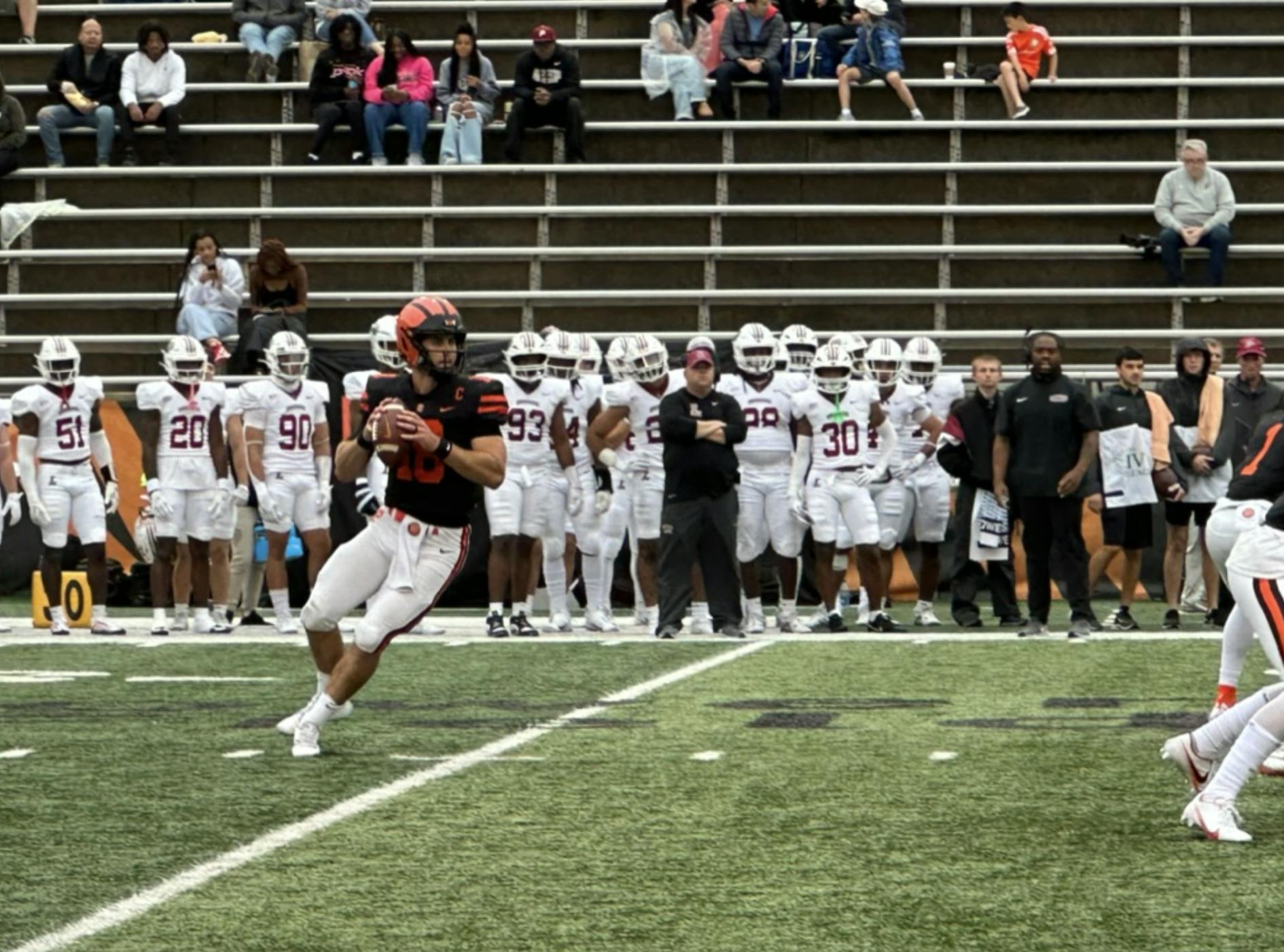 Man in black and orange holds football, looks to field, with people in white uniforms in background. 