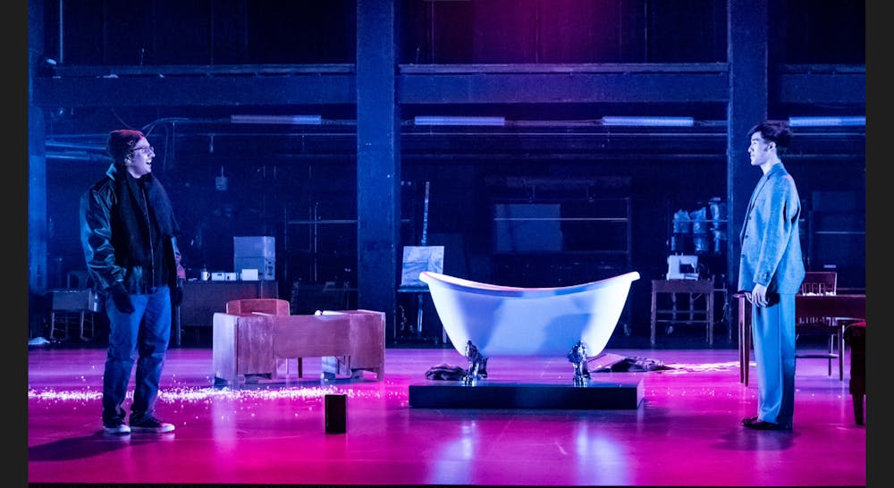Two people face each other on a stage with a bathtub and covered in purple light.