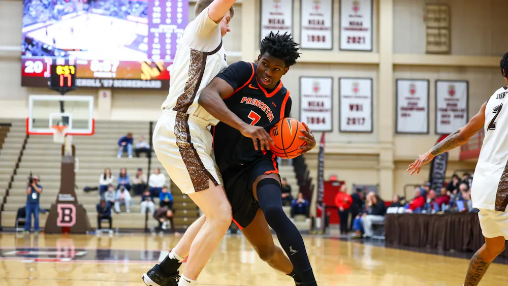 Player in a black and orange jersey drives toward the basket while being defended by a Brown player.