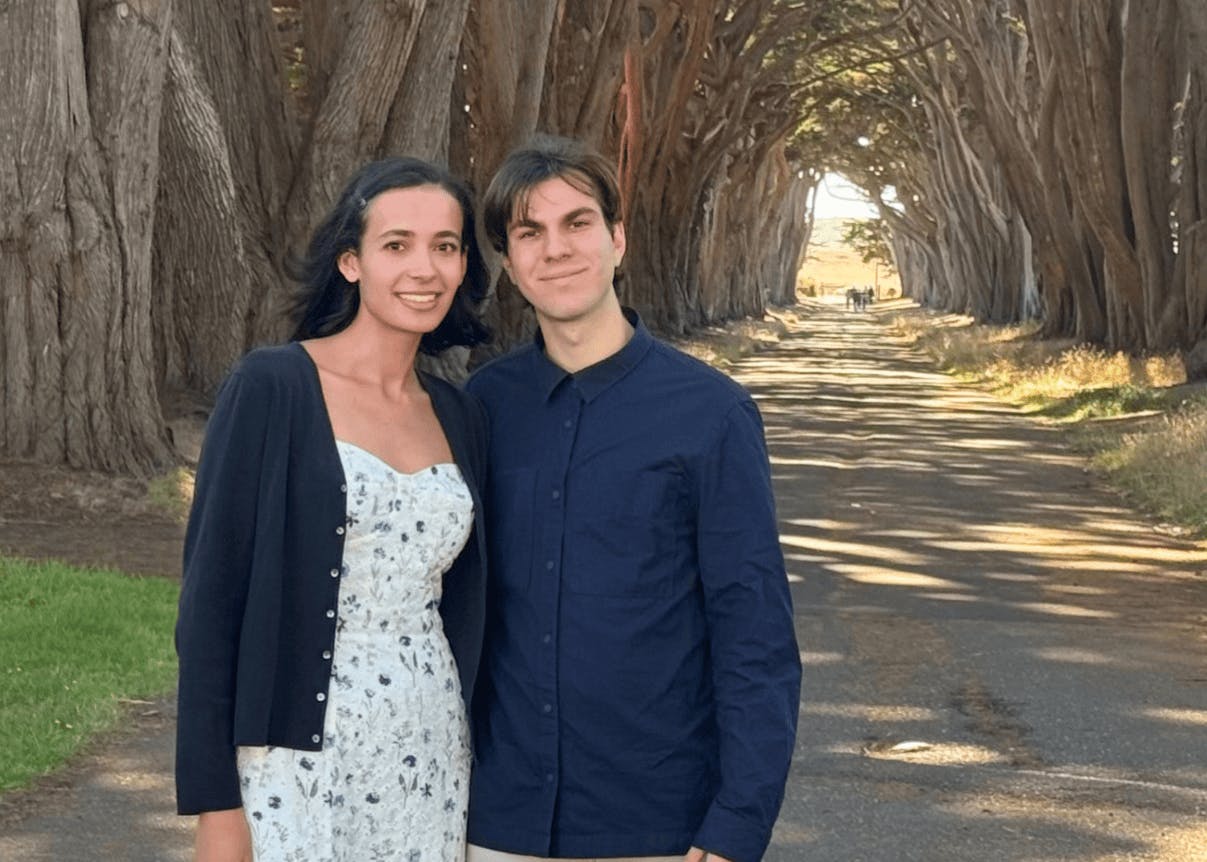 Two people standing with trees in the background.