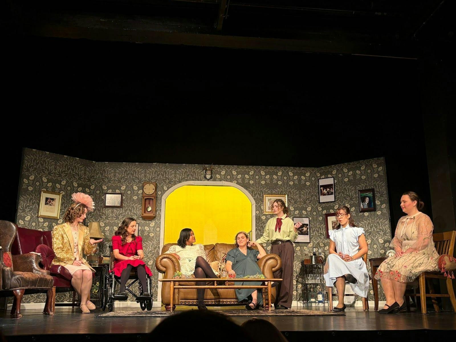 Actresses all dressed in colorful outfits of baby blue and dark red sit in various seats on a 1930s themed stage. 