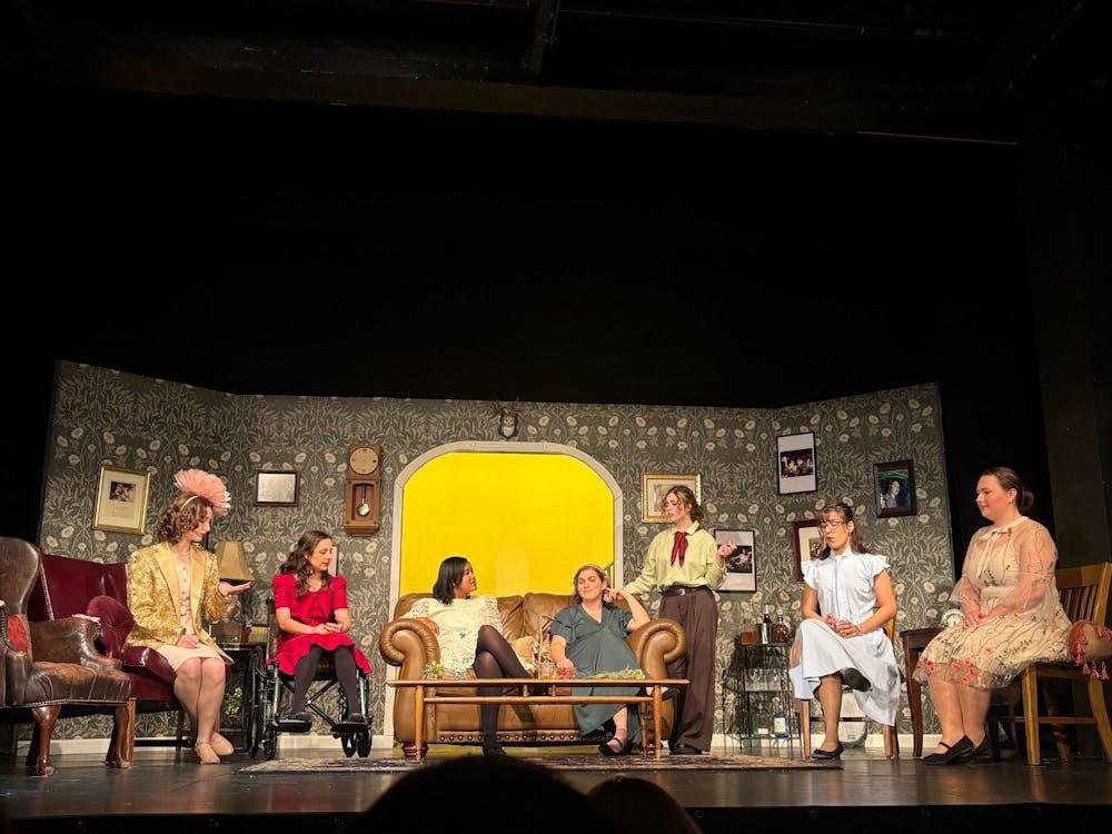 Actresses all dressed in colorful outfits of baby blue and dark red sit in various seats on a 1930s themed stage.