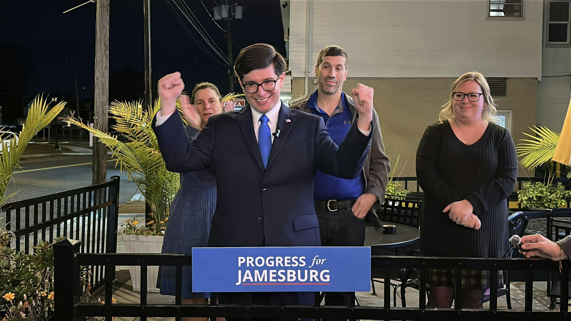 A man in a dark suit and blue tie stands behind a podium that reads “Progress for Jamesburg,” raising both fists in celebration.