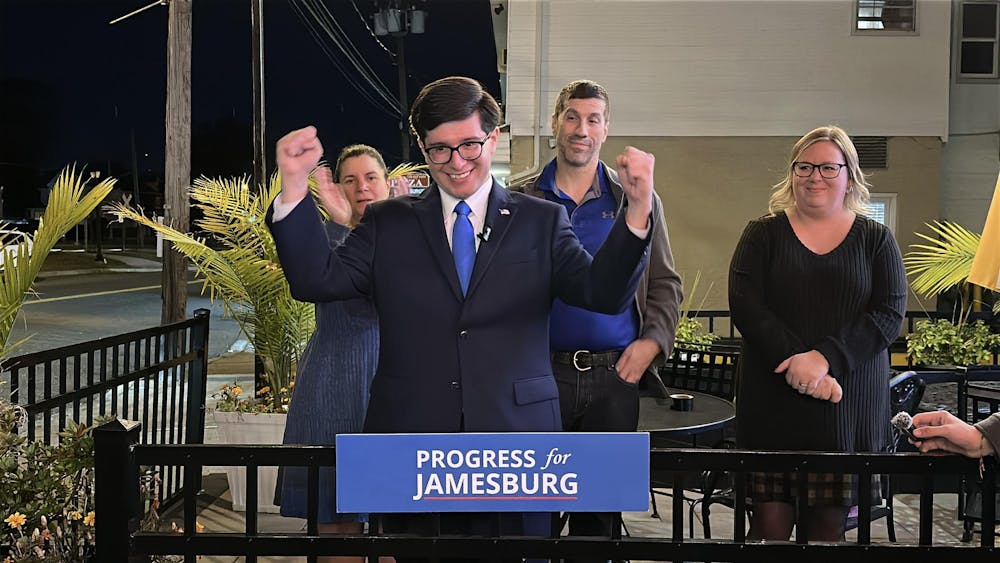 A man in a dark suit and blue tie stands behind a podium that reads “Progress for Jamesburg,” raising both fists in celebration.