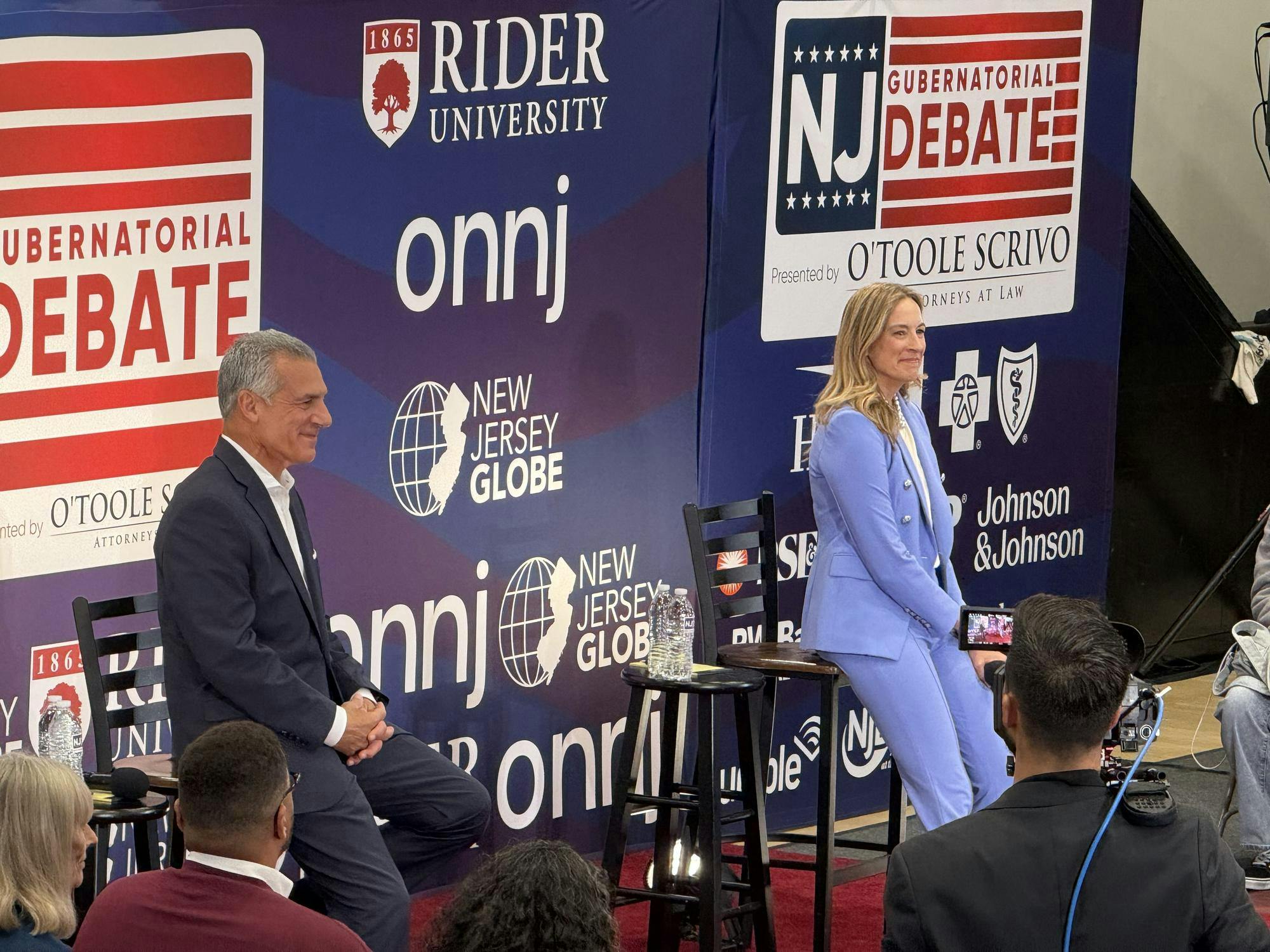 Two people sitting on chairs speaking to an audience at a politcal debate with sponsporships in the background.