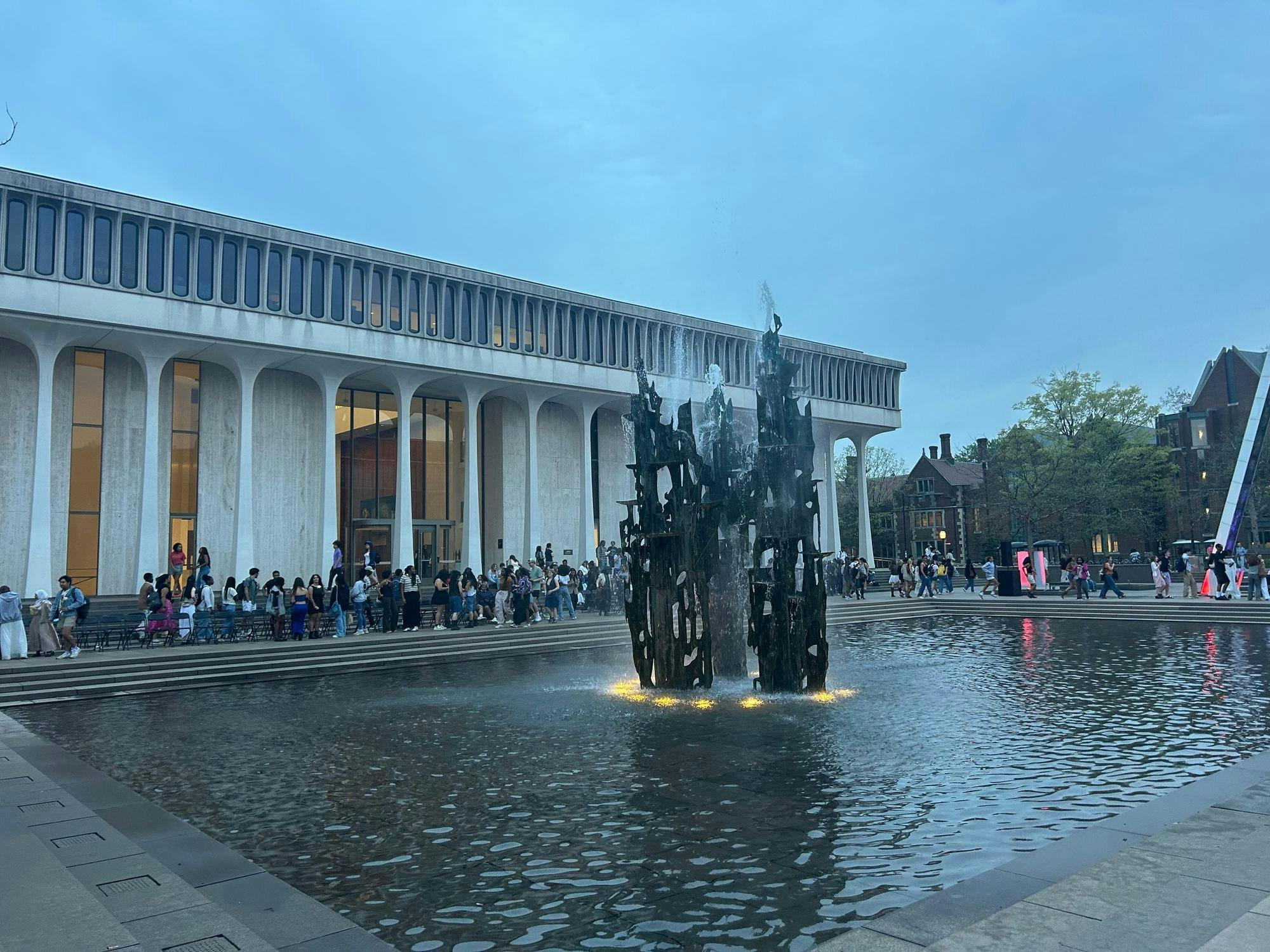 A crowd of people are near a large square fountain that is filled with water. The fountain has a tall gray abstract sculpture in it.
