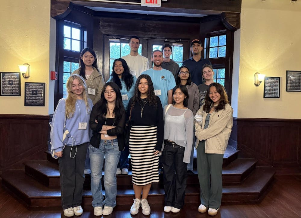 Thirteen people stand in three rows on dark wooden steps, smiling at the camera. Behind them are windows flanked by cream-colored walls.  