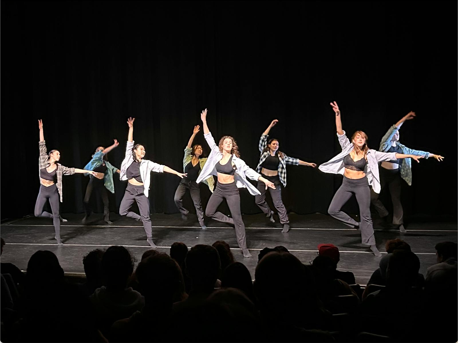 Eight dancers on stage, jumping with one arm raised.