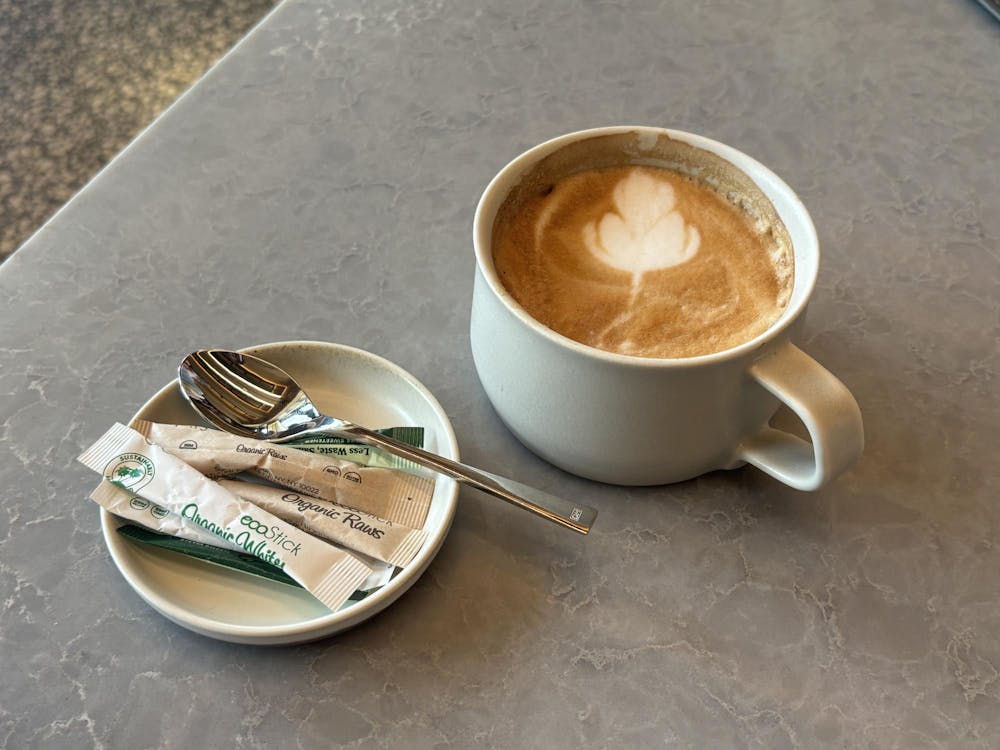 A brown latte is in a white mug. Brown and green sugar packets are on a white plate with a silver spoon resting on it.