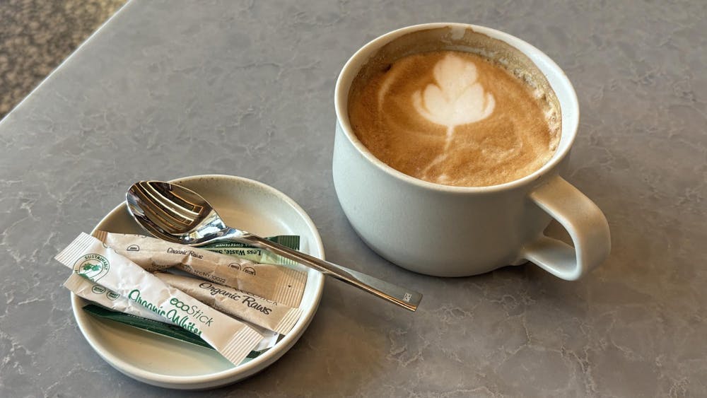 A brown latte is in a white mug. Brown and green sugar packets are on a white plate with a silver spoon resting on it.