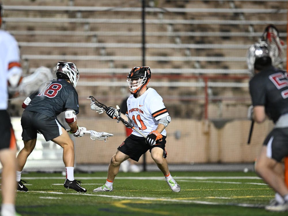 A lacrosse player wearing black shorts and a white jersey with a helmet on with a lacrosse stick in his hand matched up with a defender on the opposing team on a grass field.