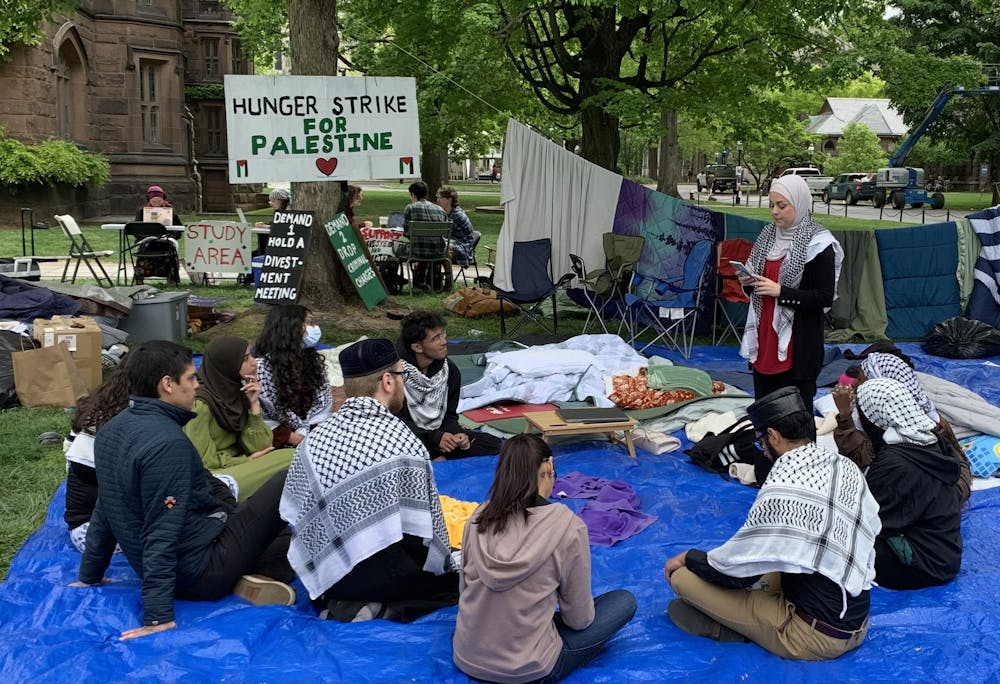 Students sit around on a blue tarp with a speaker standing. She is wearing a hijab and keffiyah and appears to be reading from her phone. In the background, there are signs saying “Hunger Strike for Palestine” and “Demand 1 Hold a Divestment Meeting”.