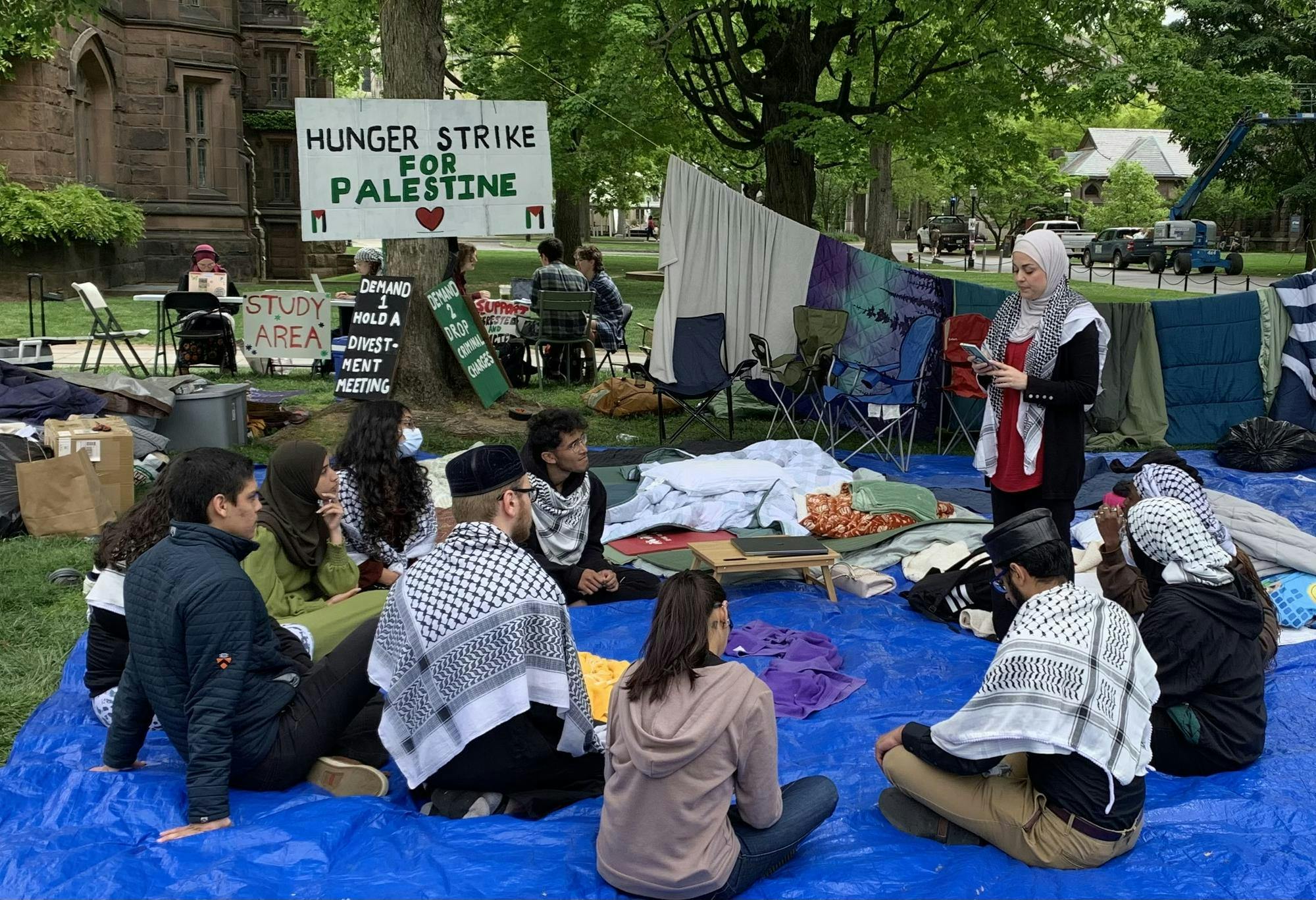 Students sit around on a blue tarp with a speaker standing. She is wearing a hijab and keffiyah and appears to be reading from her phone. In the background, there are signs saying “Hunger Strike for Palestine” and “Demand 1 Hold a Divestment Meeting”.