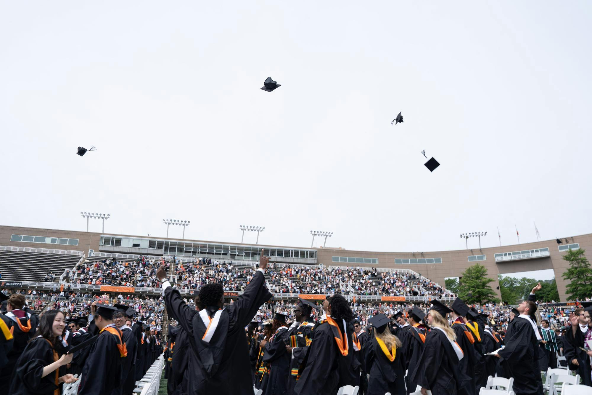 A large group of people wearing long robes throw square caps into the air.
