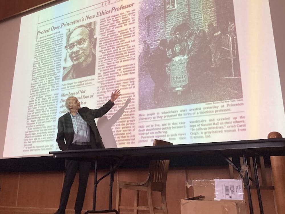 An old man gestures to a projected image of a newspaper article titled "Protest Over Princeton's New Ethics Professor"