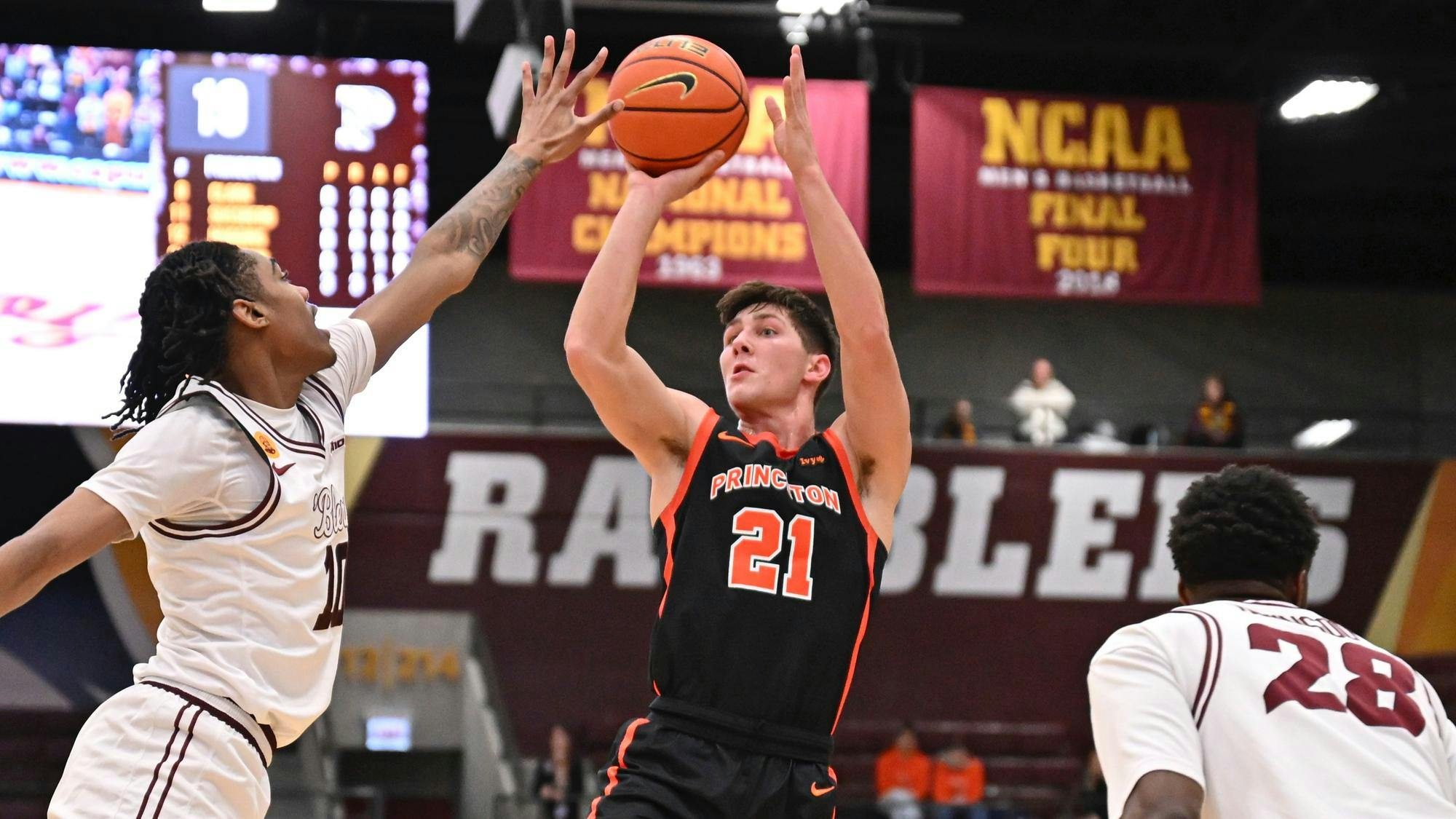 A Princeton basketball player wearing No. 21 takes a jump shot as a Loyola Chicago defender in white contests with banners in the background. 
