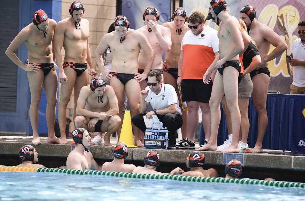 Group of men huddle around coach at poolside, with the coach talking to the six players in a pool. 