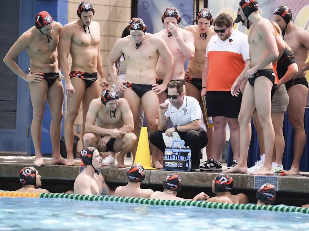 Group of men huddle around coach at poolside, with the coach talking to the six players in a pool. 