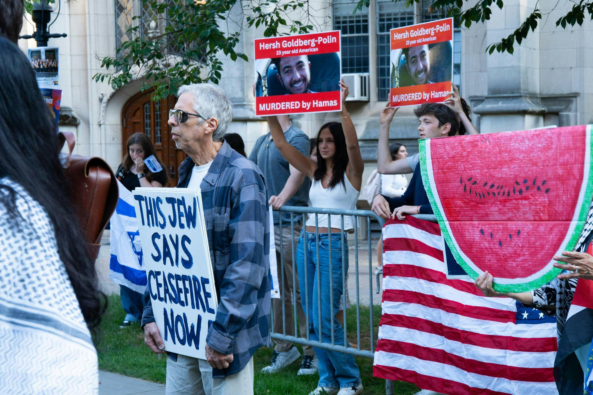 Counter-protesters stand behind a metal fence holding posters with a man's face and the American flag. The posters read: "Hersh Goldberg-Polin/27 year old American/ MURDERED by Hamas." In front of the gate, an elderly protester hold a sign that reads: "This Jew says ceasefire now." Next to him a non visible figure holds a cardboard cut out of a watermelon. 